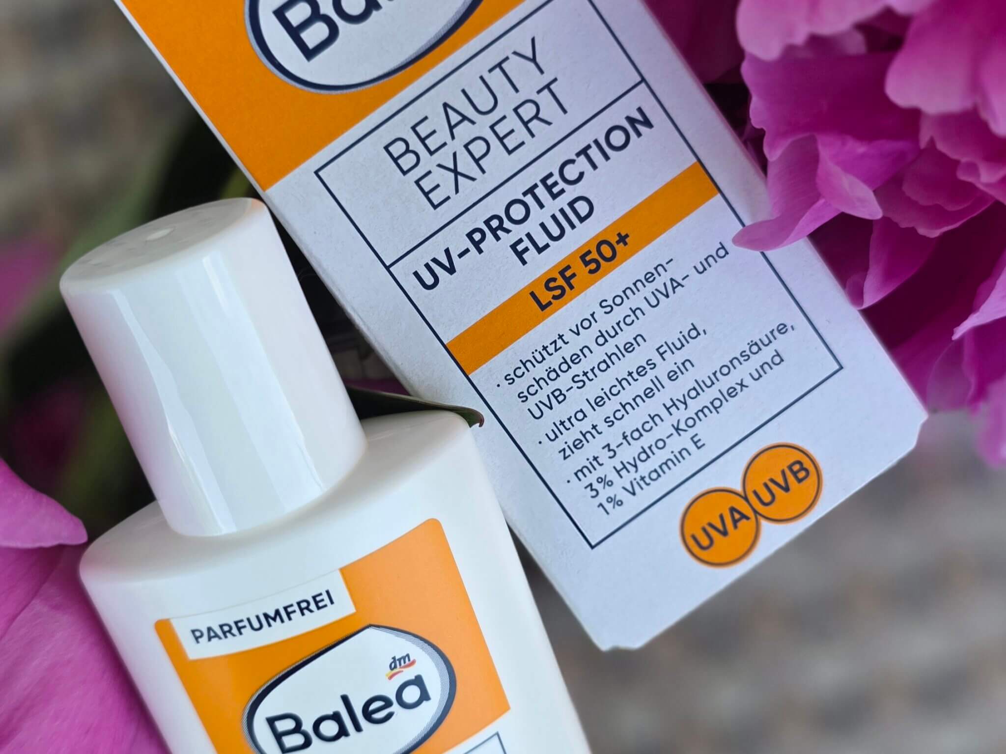 Das Balea Beauty Expert UV-Protection Fluid LSF 50+ in der handlichen weißen Flasche mit charakteristischem orange-weißem Etikett, daneben die aufgeklappte Faltschachtel mit dem Hinweis auf die verbesserte Rezeptur. Sichtbar auf der Verpackung: parfümfrei, mit Hyaluronsäure, Vitamin E und 3-fach Hydro-Komplex, Breitband-Schutz gegen UVA- und UVB-Strahlen. Das Produkt liegt zwischen üppigen rosa Pfingstrosenblüten — ein bewusst unaufgeregter Kontrast zwischen Drogerie-Pragmatismus und sommerlicher Leichtigkeit.