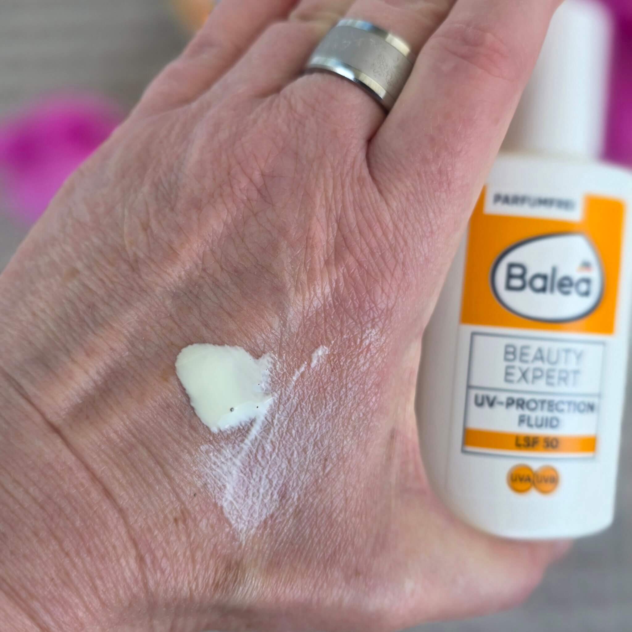 Eine reife Hand mit silbernem Ring hält die weiße Flasche des Balea Beauty Expert UV-Protection Fluid LSF 30 — auf der Handrückseite ein frischer Swatch des Fluids, dessen cremig-helle Textur bereits leicht einzieht. Die Konsistenz wirkt leicht und geschmeidig, nicht schwer oder fettig. Im Hintergrund unscharf: rosa Blüten, graues Holz. Das Bild zeigt, wie fein das parfümfreie Sonnenschutz-Fluid auf der Haut liegt — ideal für einen ersten Eindruck von Textur und Verträglichkeit.
