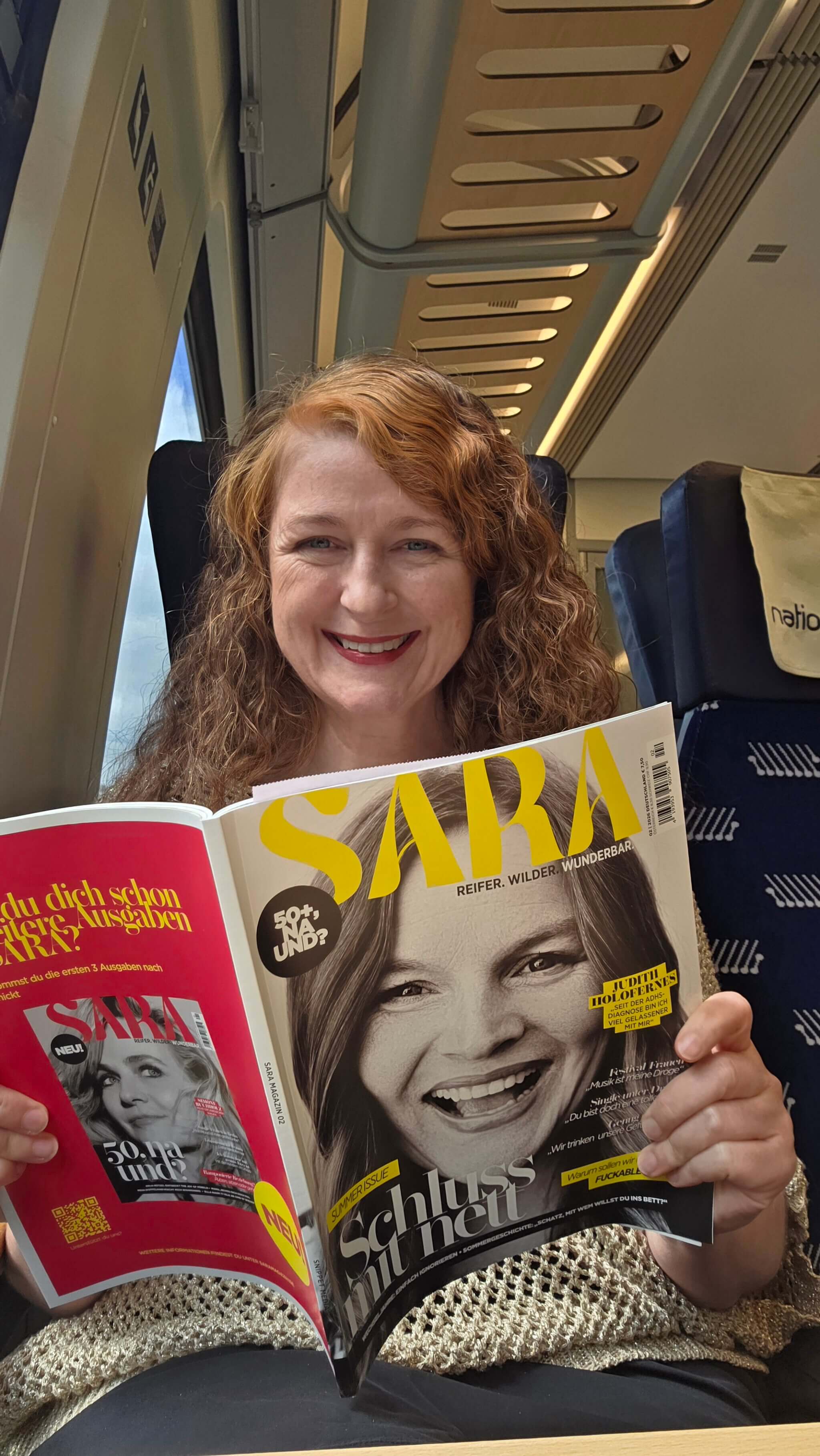 Cerstin von stylerebelles im Zug mit der ersten Ausgabe des Sara Magazins in den Händen, Cover mit dem Schriftzug "Reifer. Wilder. Wunderbar." und dem Slogan "50+, na und?". Sie trägt einen goldschimmernden Strickpullover, ihre roten Locken fallen offen über die Schultern, dezenter roter Lippenstift. Im Hintergrund das Fenster eines ICE mit vorbeiziehender Landschaft. Eine Szene, die Lesefreude und Leichtigkeit einfängt – die neue Frauenzeitschrift für Frauen über 50 unterwegs gelesen.